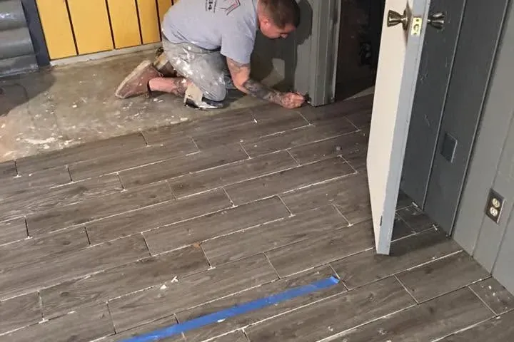 Worker kneeling installing gray wood-look plank tile in room, C&A Roofing & Construction