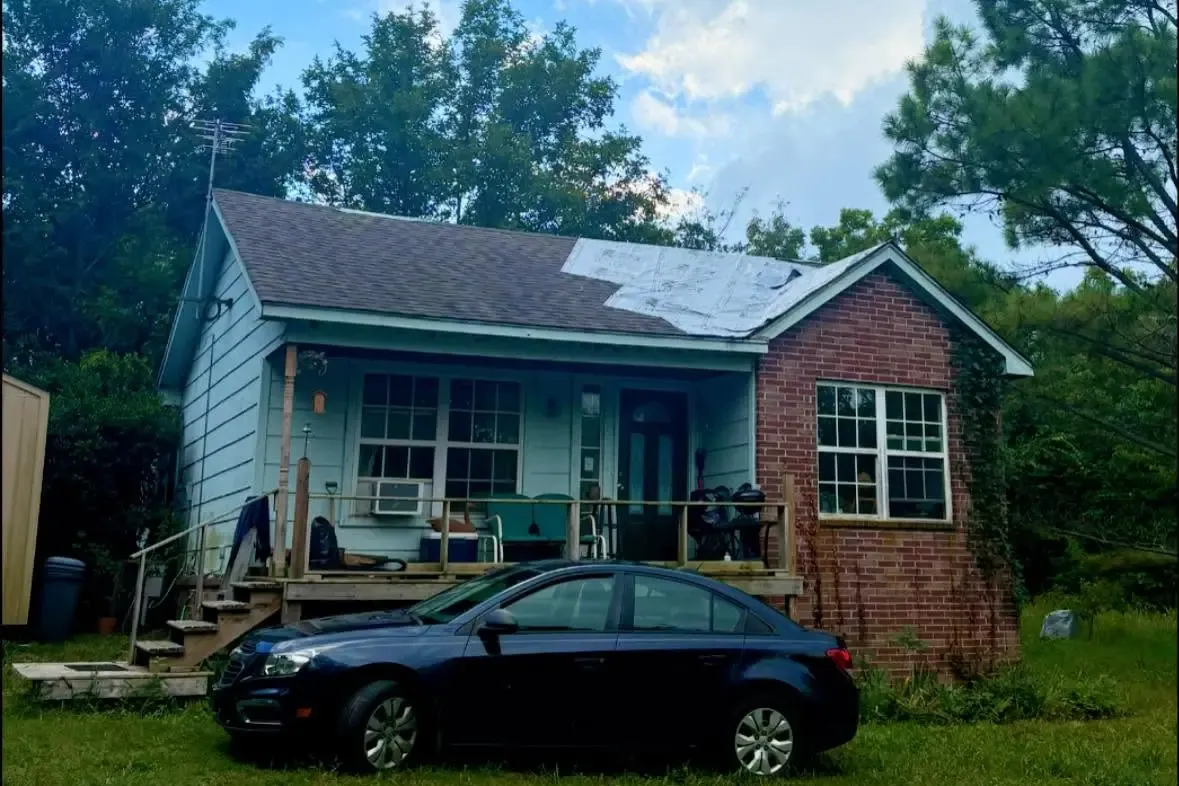 Blue house with partially tarped/damaged roof, car in front, C&A Roofing & Construction