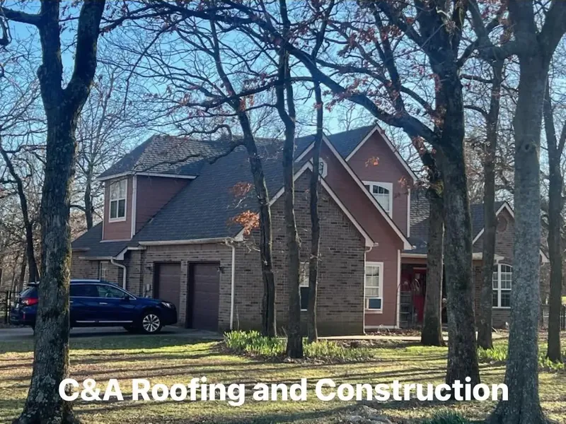 Two-story brick house with dark shingle roof among trees, C&A Roofing & Construction